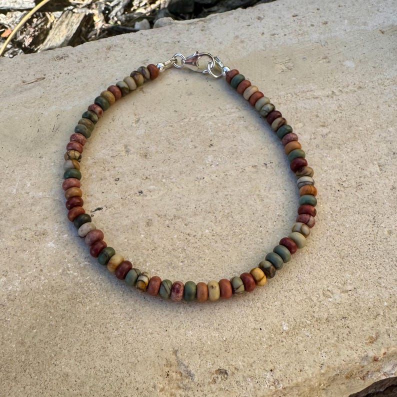 May include: A beaded bracelet featuring a variety of earth-toned beads in shades of brown, green, and red. The bracelet is secured with a silver clasp. The beads are small and round, creating a delicate and stylish accessory.