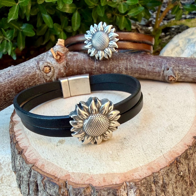 May include: Two leather bracelets with sunflower accents. One bracelet is black, the other is brown. Both bracelets have a silver-colored sunflower in the center. The bracelets are displayed on a wooden surface.