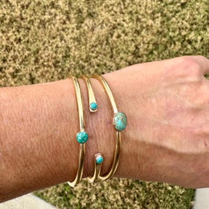 May include: Gold-coloured metal bracelets with turquoise stones. The bracelets have a simple, elegant design, with the turquoise stones adding a pop of colour. The bracelets are worn on a wrist.