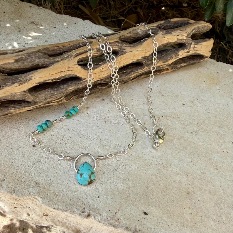 May include: A silver chain necklace featuring a teardrop-shaped turquoise pendant suspended from a small silver ring. The necklace includes a series of small, round turquoise beads integrated into one side of the silver link chain.