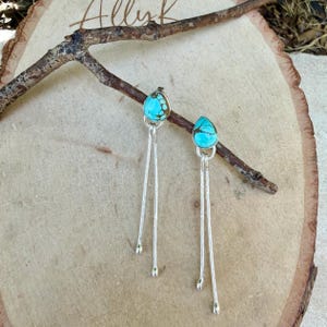 May include: A pair of dangle earrings featuring teardrop-shaped turquoise stones with dark matrix patterns, set in silver bezels. Each earring has two long, thin, textured silver stems extending downward from the stone, creating a linear design.