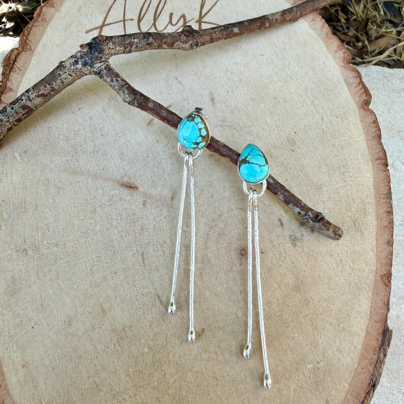 May include: A pair of dangle earrings featuring teardrop-shaped turquoise stones with dark matrix patterns, set in silver bezels. Each earring has two long, thin, textured silver stems extending downward from the stone, creating a linear design.