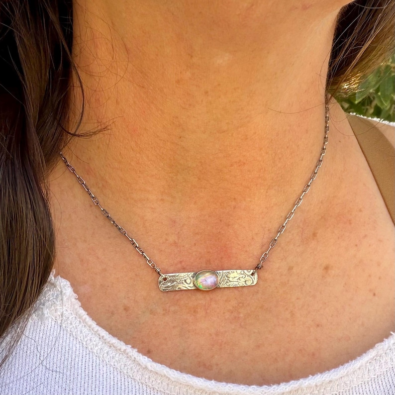 May include: A silver-toned necklace featuring a rectangular metal bar pendant with an etched pattern and a central, iridescent, oval-shaped cabochon stone. The pendant is suspended from a delicate, thin silver-toned metal chain.