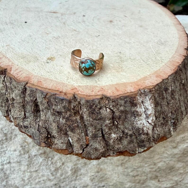 May include: A copper-colored adjustable ring with a round, turquoise-colored stone. The ring has a textured band and is set on a wooden surface. The ring is a piece of jewelry.