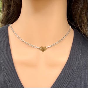 May include: A silver-toned chain necklace featuring a central, small, hammered-texture gold-toned heart charm. The chain consists of oval-shaped links connected to the heart pendant with wire-wrapped details, resting against a dark ribbed fabric.