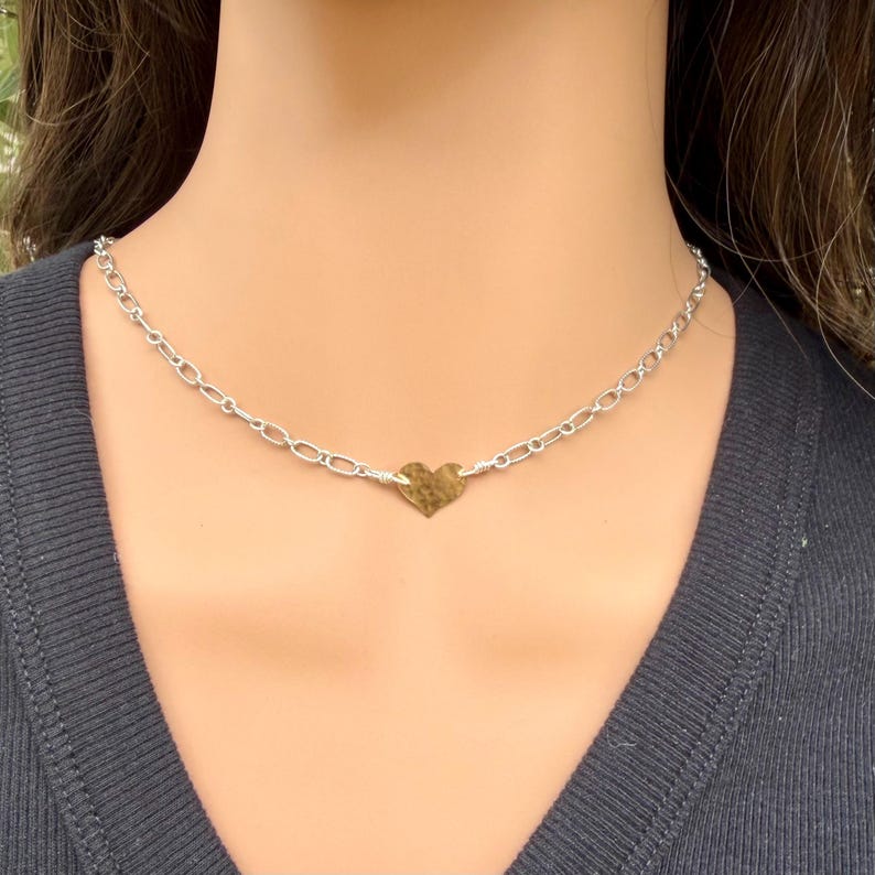 May include: A silver-toned chain necklace featuring a central, small, hammered-texture gold-toned heart charm. The chain consists of oval-shaped links connected to the heart pendant with wire-wrapped details, resting against a dark ribbed fabric.