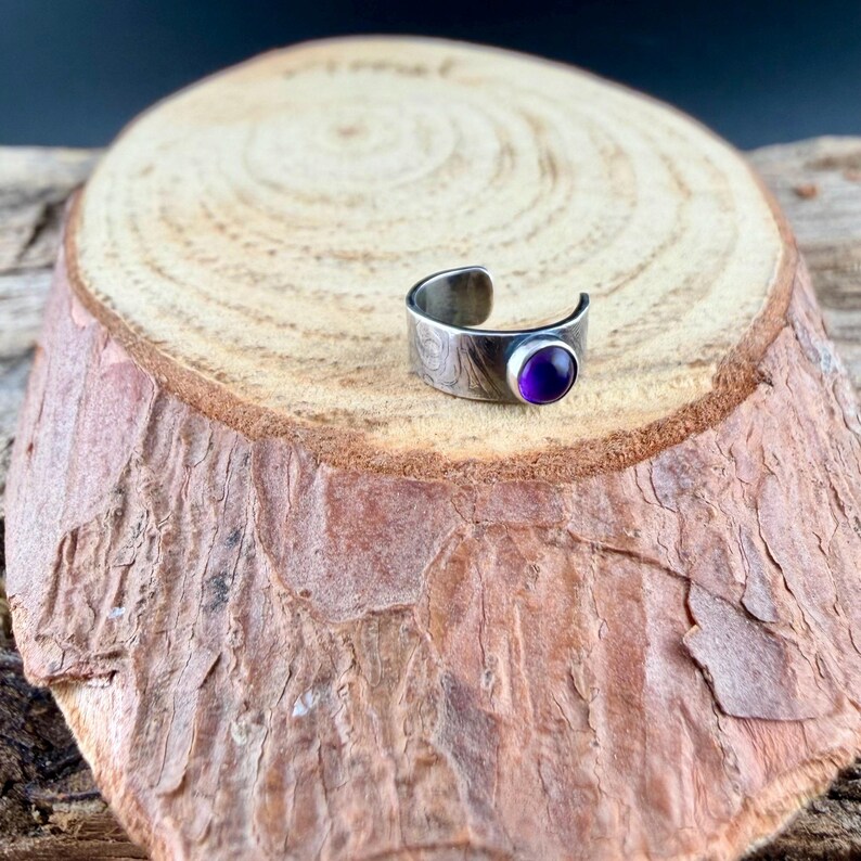 May include: A silver-toned adjustable ring with an engraved pattern and a round, deep purple gemstone. The ring is set on a natural wood slice, showcasing its design and color.