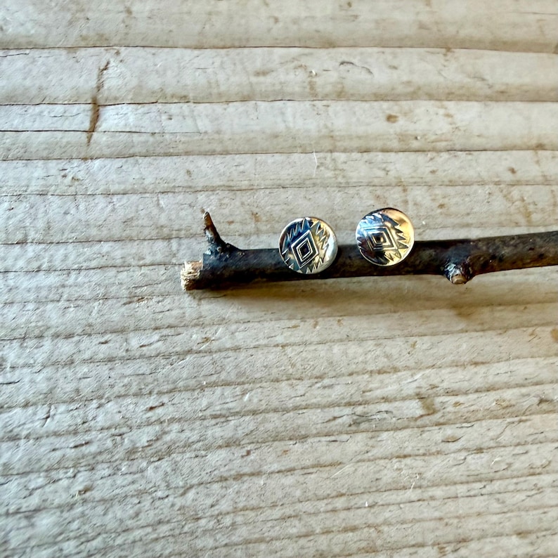 May include: Two circular silver stud earrings featuring a stamped geometric diamond pattern in the center, surrounded by small radiating lines. The earrings are resting on a dark brown twig.