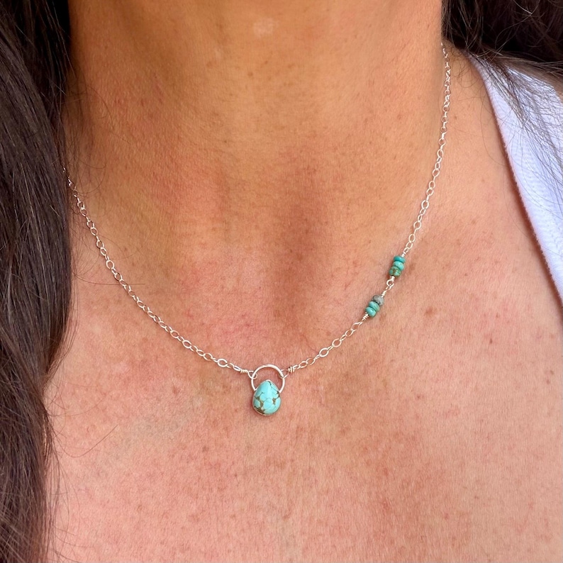 May include: A delicate silver chain necklace featuring a single teardrop-shaped turquoise pendant suspended from a small circular silver loop. The chain includes two additional small turquoise beads positioned asymmetrically along the right side.