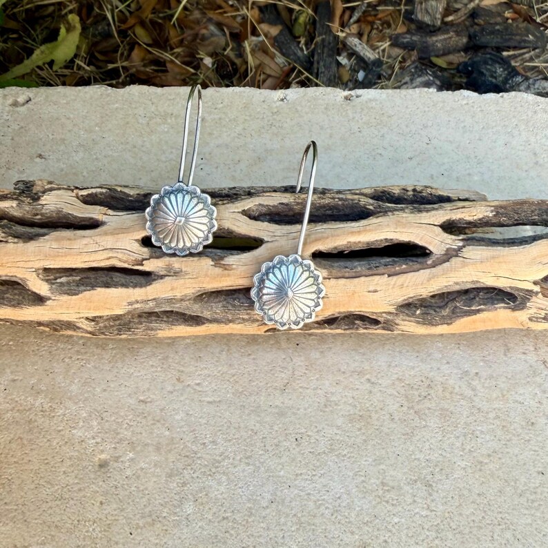 May include: A pair of silver-toned drop earrings featuring a circular, sunburst-inspired design with radiating lines and a central point. Each earring is attached to a curved wire hook, showcasing a metallic finish with intricate, symmetrical detailing.