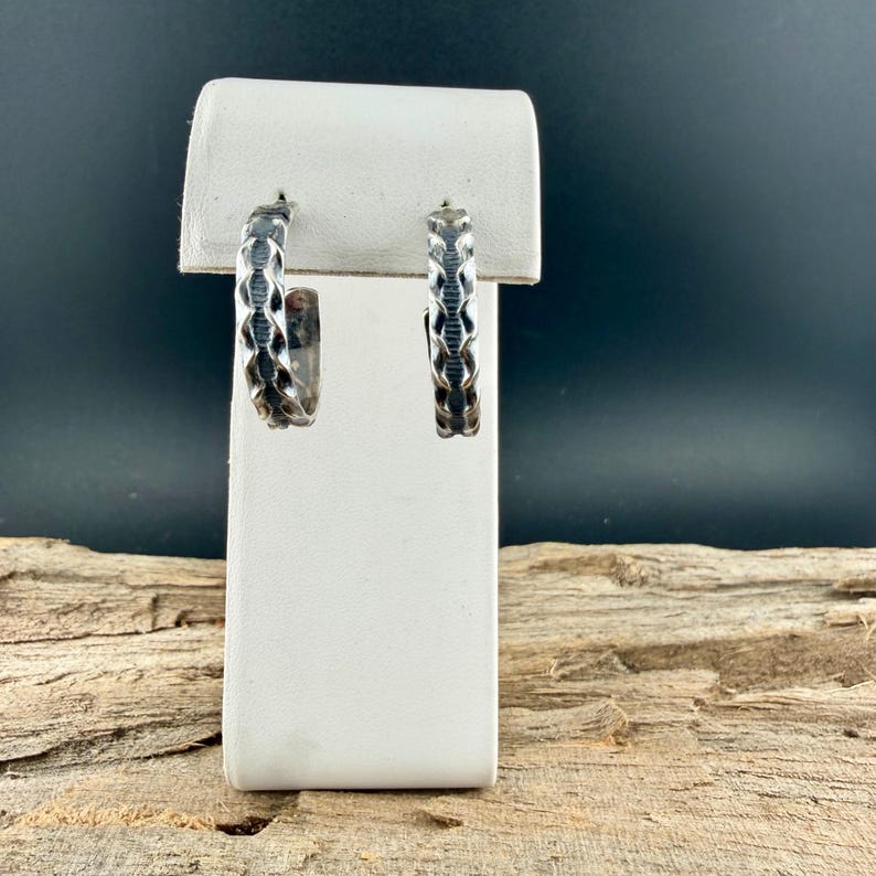 May include: A pair of silver hoop earrings with a textured, geometric design. The earrings are displayed on a white stand against a dark background. The earrings have a repeating pattern of connected shapes.