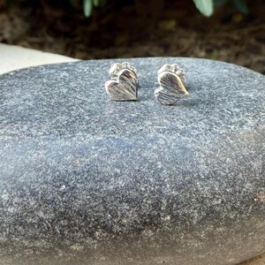 May include: A pair of silver-toned stud earrings shaped like hearts. Each heart features a textured, grooved surface finish and a metallic sheen. The earrings are small, compact, and designed with a post-style backing for secure attachment.