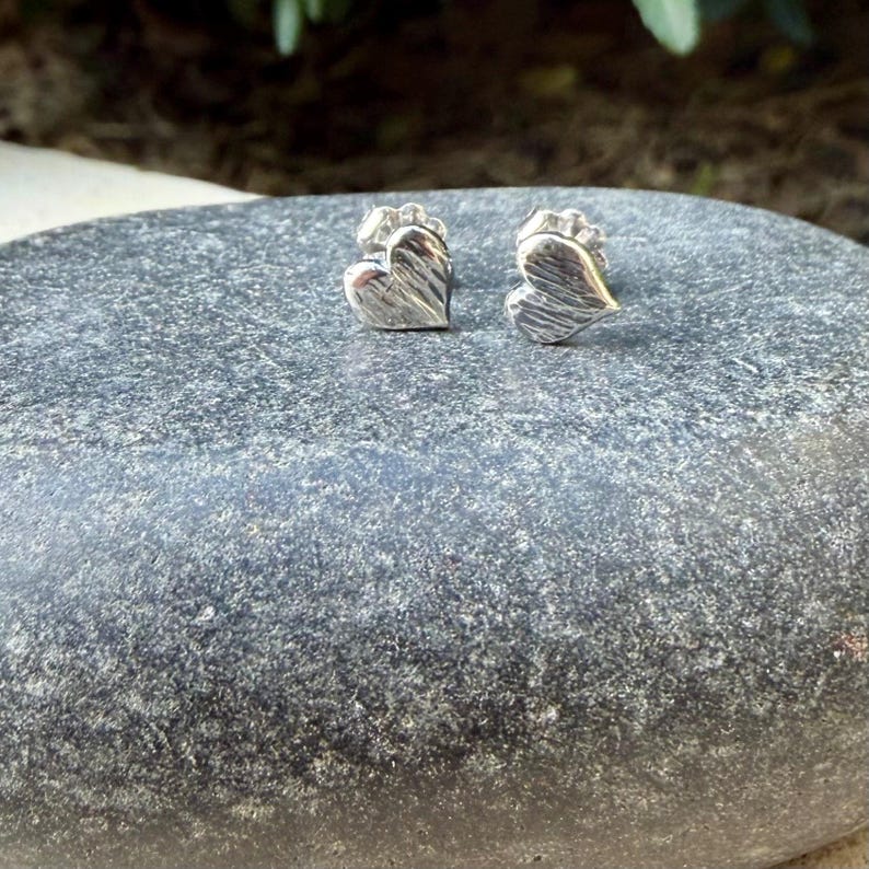 May include: A pair of silver-toned stud earrings shaped like hearts. Each heart features a textured, grooved surface finish and a metallic sheen. The earrings are small, compact, and designed with a post-style backing for secure attachment.