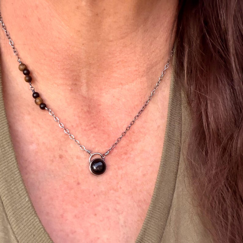 May include: A silver-toned chain necklace featuring a single round black stone pendant held by a wire-wrapped bail. The chain includes an asymmetrical accent section with alternating small brown tiger eye and black beads on one side of the necklace.