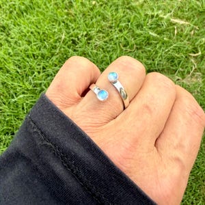 May include: A silver ring featuring two round, pale blue gemstones. The open design ring has one stone on top of the finger and the other on the side. The ring is on a hand, with a black sleeve, against a backdrop of green grass.