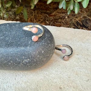 May include: Two open-style rings crafted from silver-toned metal, each featuring two small, round, pale pink cabochon stones set at the ends of the band. The rings have a textured, hammered finish along the metal surface, creating a reflective appearance.