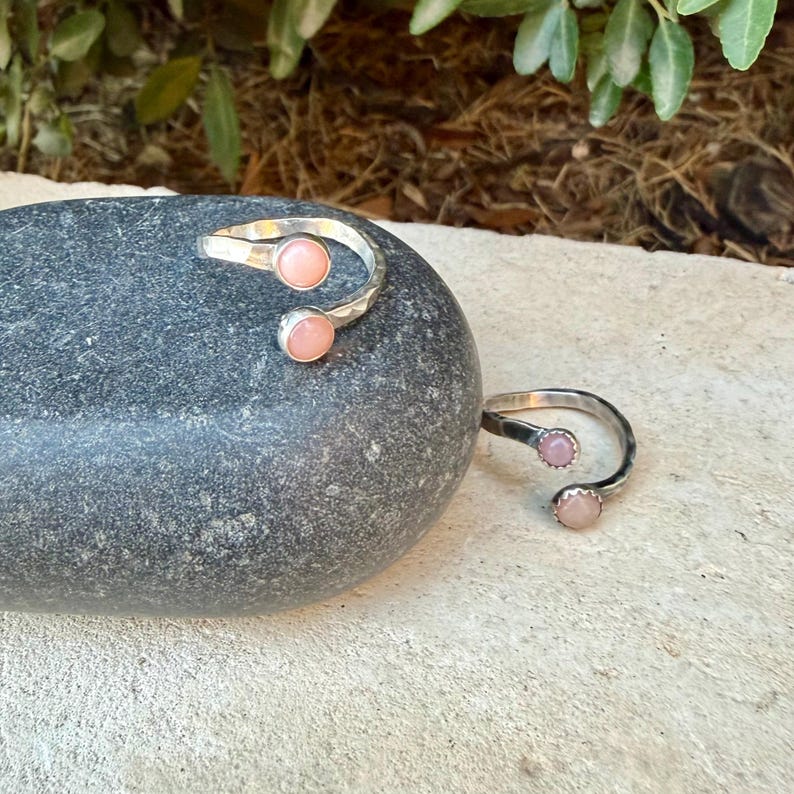 May include: Two open-style rings crafted from silver-toned metal, each featuring two small, round, pale pink cabochon stones set at the ends of the band. The rings have a textured, hammered finish along the metal surface, creating a reflective appearance.