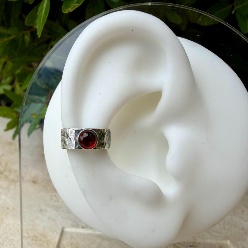May include: A silver ear cuff with a textured band and a round, deep red gemstone. The cuff is displayed on a white ear model against a backdrop of green foliage.