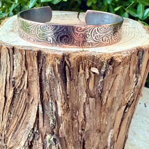 Puede incluir: Brazalete de pu&ntilde;o de cobre con un intrincado dise&ntilde;o de remolinos. El dise&ntilde;o de pu&ntilde;o abierto presenta un dise&ntilde;o detallado en relieve en la parte delantera. El brazalete se exhibe sobre una superficie de madera r&uacute;stica, mostrando su artesan&iacute;a.