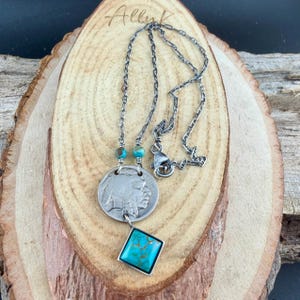 Op de afbeelding: Zilverkleurige ketting met een munt-hanger met een Indiaans profiel en een vierkante turkoois steen. De ketting heeft een ketting met turkoois kralen en een karabijnsluiting. De ketting wordt gepresenteerd op een houten oppervlak.