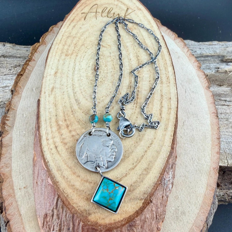 Op de afbeelding: Zilverkleurige ketting met een munt-hanger met een Indiaans profiel en een vierkante turkoois steen. De ketting heeft een ketting met turkoois kralen en een karabijnsluiting. De ketting wordt gepresenteerd op een houten oppervlak.