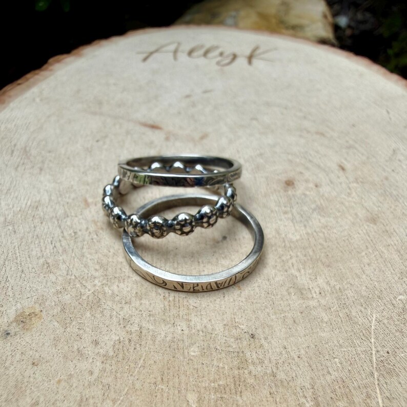 May include: Three silver rings stacked on a wooden surface. The top ring has small, evenly spaced details. The middle ring features a floral design. The bottom ring has etched details. The word "Allyk" is etched on the wood.