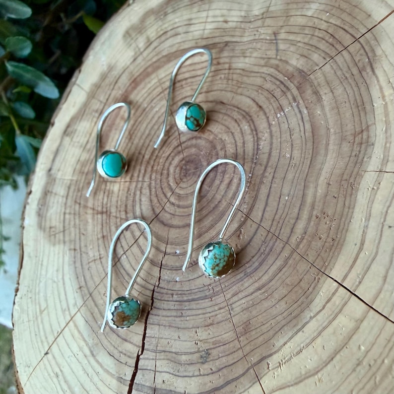 May include: Four silver drop earrings featuring small, round turquoise stones with brown matrix patterns. Each earring has a simple, curved silver wire hook and a polished metal setting that secures the circular gemstone in place.