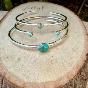 May include: Three silver-toned cuff bracelets with turquoise stones. The bracelets are stacked on a light brown wooden surface. The turquoise stones are round and set in a bezel setting. The bracelets are simple and elegant.