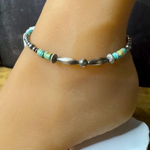 May include: Anklet featuring turquoise and silver beads. The anklet has a mix of small, round silver beads, elongated silver beads, and turquoise-colored beads. The anklet is worn on the ankle.
