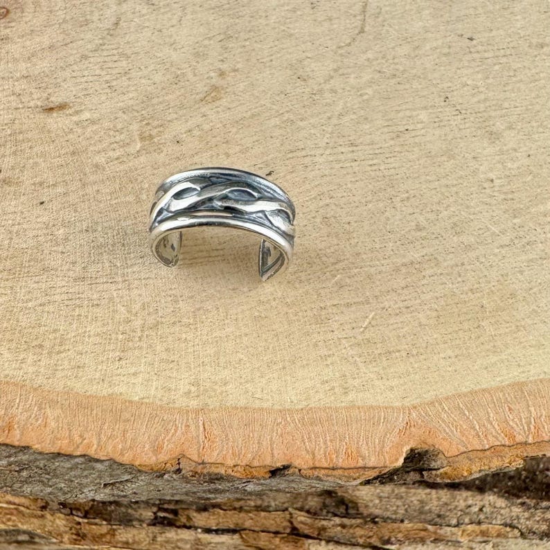 May include: A silver-colored ring with an open design. The ring features a textured, wave-like pattern around the band. The ring is displayed on a light brown wooden surface.