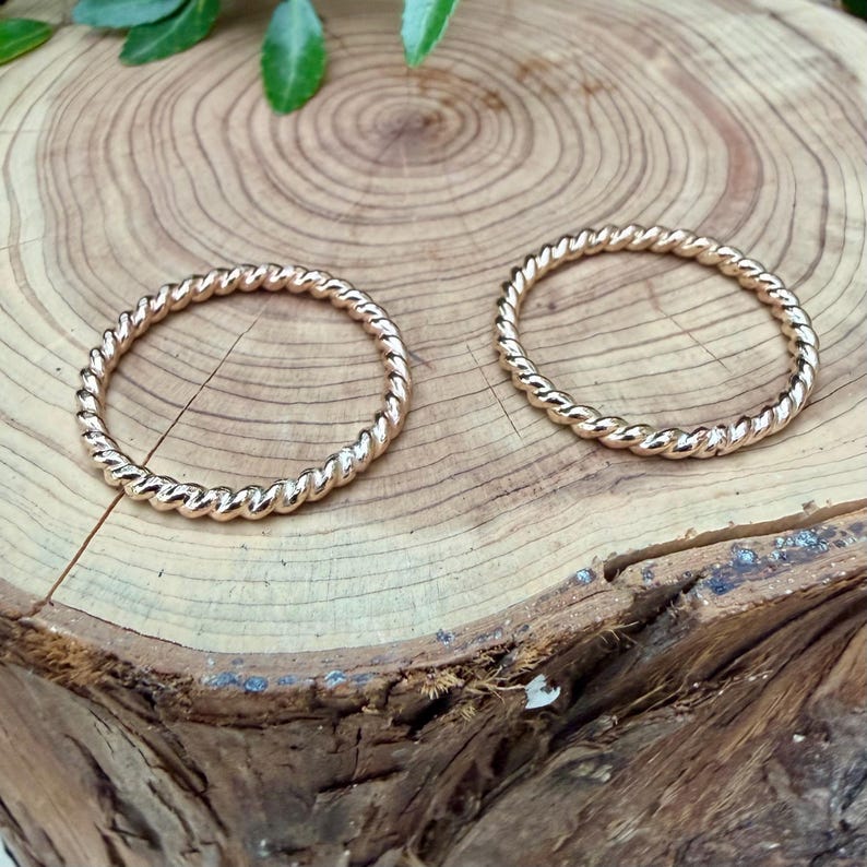 May include: Two gold-colored, twisted rope design rings. The rings are circular and sit on a light brown, textured wooden surface. The rings are made of a shiny metal and have a detailed, braided appearance. The background includes green leaves.