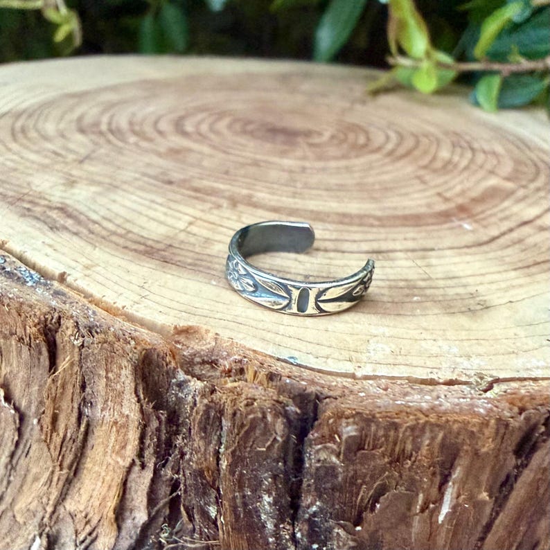 K&ouml;nnte beinhalten: Ein silberfarbener offener Ring mit eingraviertem Blumenmuster, Blattmotiven und einem ovalen Detail in der Mitte. Das Metall weist in den vertieften Bereichen des Designs ein dunkles, oxidiertes Finish auf, das die botanischen Gravuren betont.