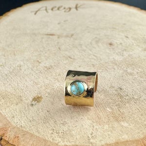 May include: A wide, gold-colored ring with a hammered texture. The ring features a round, light blue turquoise stone set in the center. The ring is displayed on a wooden surface.