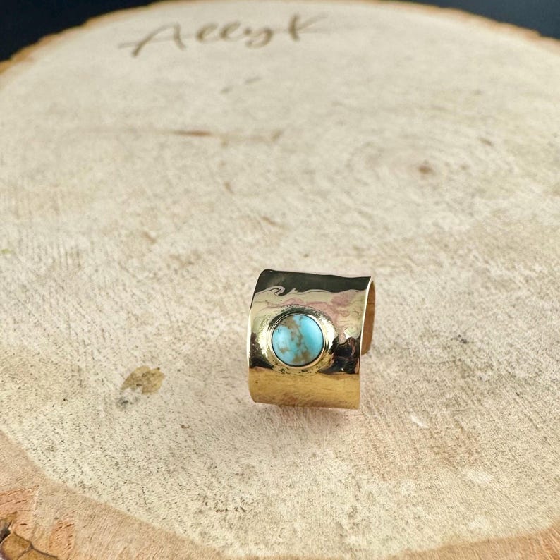 May include: A wide, gold-colored ring with a hammered texture. The ring features a round, light blue turquoise stone set in the center. The ring is displayed on a wooden surface.