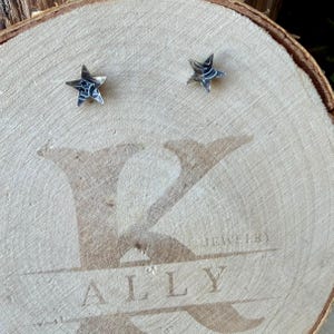 May include: A pair of silver star-shaped stud earrings with a dark, oxidized finish and etched detailing. The earrings are displayed on a circular wooden slice featuring the engraved text K ALLY JEWELRY across the center surface.