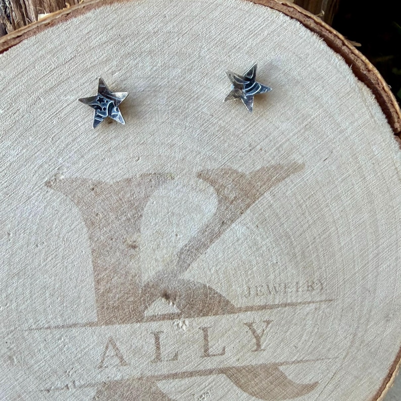 May include: A pair of silver star-shaped stud earrings with a dark, oxidized finish and etched detailing. The earrings are displayed on a circular wooden slice featuring the engraved text K ALLY JEWELRY across the center surface.
