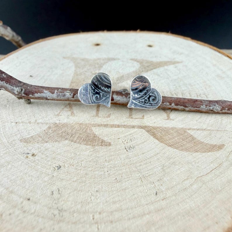 May include: A pair of heart-shaped stud earrings crafted from silver-toned metal. Each earring features a detailed, dark-toned engraved pattern across its surface. The earrings are positioned side-by-side, resting on a thin, horizontal wooden branch.