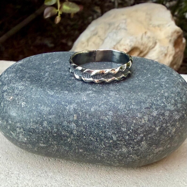 May include: A silver-toned metal ring featuring a band with a textured, wavy edge design and a central horizontal grooved pattern. The ring has a polished finish and a consistent width throughout the circumference of the circular band.
