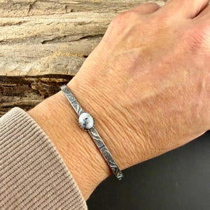 May include: A silver cuff bracelet featuring a central, round white stone with black speckles. The bracelet has an etched design. The bracelet is approximately 15 cm in circumference.