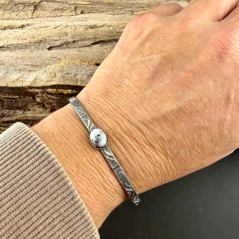 May include: A silver cuff bracelet featuring a central, round white stone with black speckles. The bracelet has an etched design. The bracelet is approximately 15 cm in circumference.