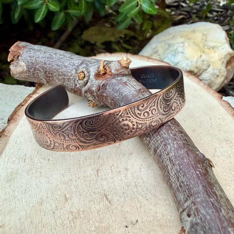 Puede incluir: Un brazalete de cobre con un intrincado dise&ntilde;o en espiral. El brazalete tiene un interior oscuro y descansa sobre una rama de madera. Las palabras "GENUINE COPPER" son visibles en el interior del brazalete.
