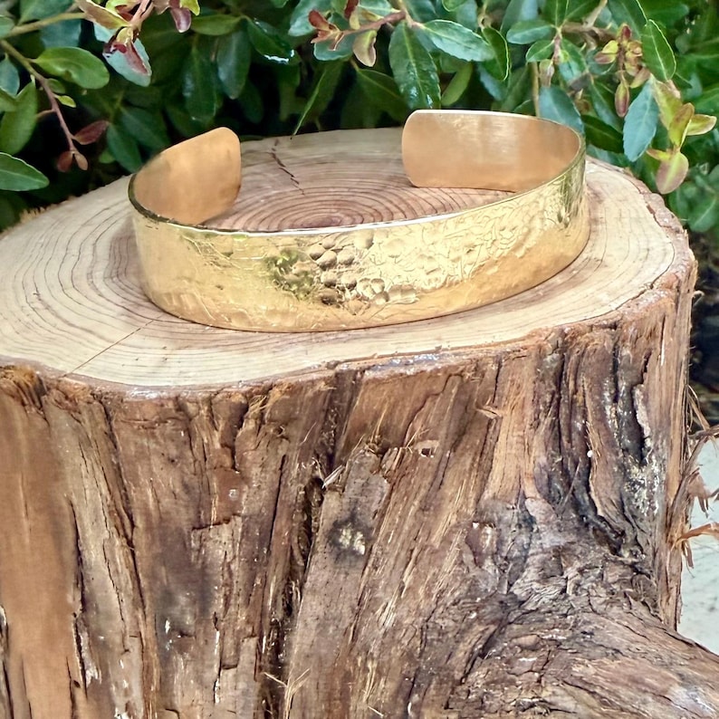 Puede incluir: Una pulsera tipo brazalete ancha y abierta, fabricada en metal pulido de tono dorado. La superficie exterior presenta una textura martillada distintiva, creando un acabado reflectante e irregular en toda la banda. Tiene un dise&ntilde;o en forma de C.