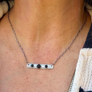 Pu&ograve; includere: Una collana color argento con un ciondolo a barra rettangolare caratterizzato da un motivo geometrico testurizzato. Il ciondolo &egrave; incastonato con due piccole pietre nere rotonde ai lati e una pietra nera quadrata al centro.