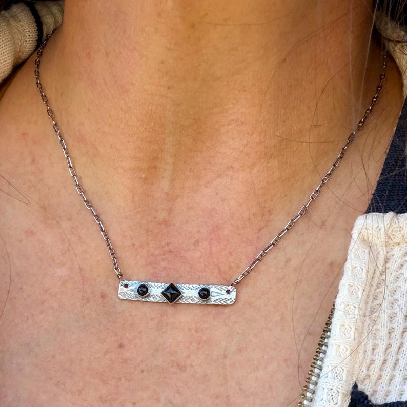 Pu&ograve; includere: Una collana color argento con un ciondolo a barra rettangolare caratterizzato da un motivo geometrico testurizzato. Il ciondolo &egrave; incastonato con due piccole pietre nere rotonde ai lati e una pietra nera quadrata al centro.