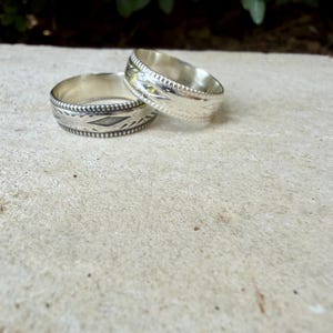 May include: Two silver rings featuring a polished finish and intricate engraved patterns along the bands. The rings have a beaded edge detail and a smooth interior surface, showcasing a metallic luster and detailed craftsmanship on the exterior.