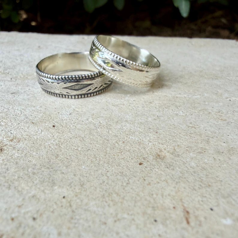 May include: Two silver rings featuring a polished finish and intricate engraved patterns along the bands. The rings have a beaded edge detail and a smooth interior surface, showcasing a metallic luster and detailed craftsmanship on the exterior.