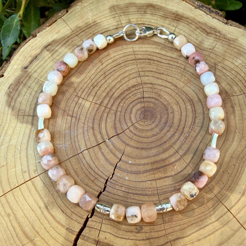 May include: A beaded bracelet featuring faceted, cube-shaped stones in shades of pale pink, cream, and soft peach. The bracelet is accented with small, cylindrical silver-toned metal spacers and finished with a silver-toned metal lobster claw clasp.