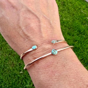 May include: A gold-toned cuff bracelet with three turquoise accents. The bracelet has an open design, with a turquoise stone at the center and two smaller turquoise accents at the ends. The bracelet is worn on a wrist.