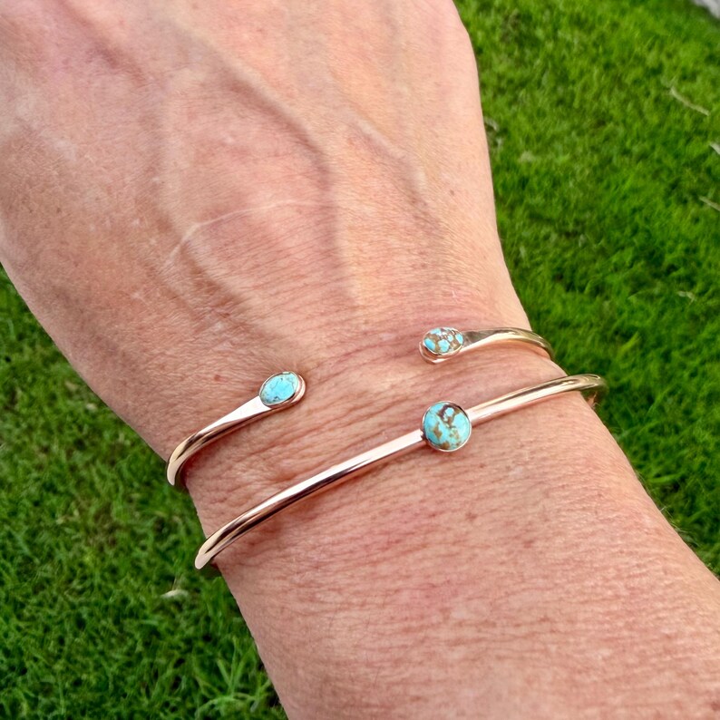 May include: A gold-toned cuff bracelet with three turquoise accents. The bracelet has an open design, with a turquoise stone at the center and two smaller turquoise accents at the ends. The bracelet is worn on a wrist.