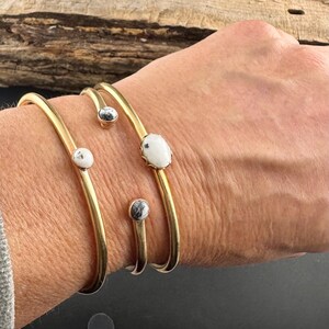 May include: Gold-toned metal bracelets with white and gray stone accents. The bracelets have a simple, elegant design, with the stones set in a minimalist style. The bracelets are shown on a person's wrist.